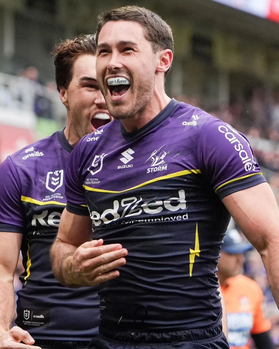 Two Melbourne Storm players celebrate on the field, one grinning in his custom mouthguard.